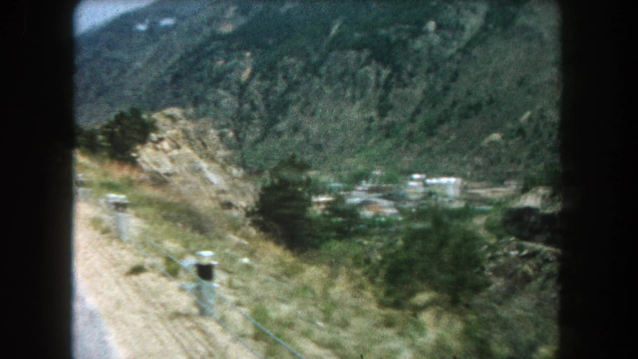1962 8mm view from a moving car on a Summit County, Colorado mountain road with roadside posts, slopes, and valley buildings.