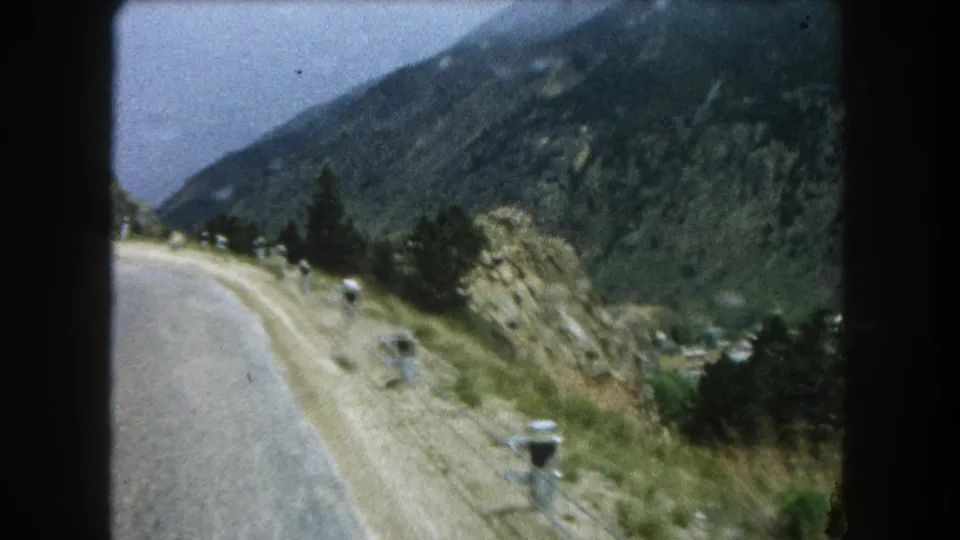 A 1962 home-movie frame from Summit County, Colorado showing a curving road, white posts, rocky hillside, and mountain backdrop.