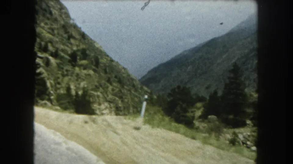 Summit County mountain-road view with a pale road shoulder, pines, and steep green terrain.