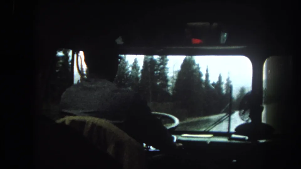 Inside a 1962 tour bus in Summit County, the driver is seen from behind while the windshield frames trees and roadway.