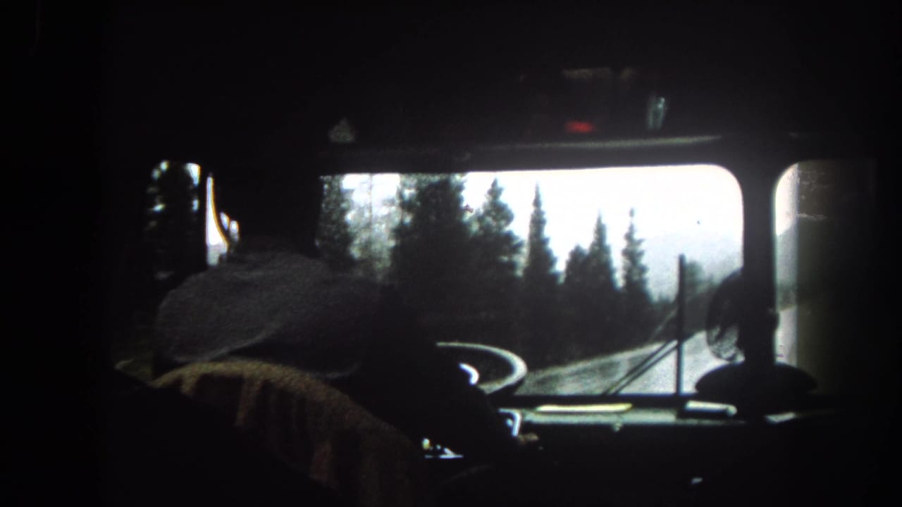 1962 Summit County, Colorado 8mm frame showing a driver silhouette, steering wheel, dashboard, and curving snowy forest road ahead.