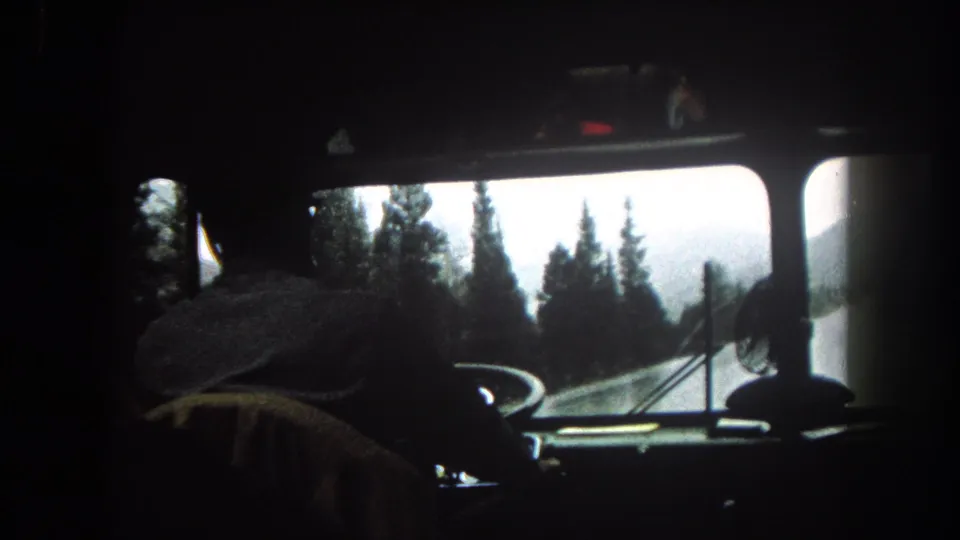 1962 Colorado home-movie frame with a shadowed driver, dashboard details, and snow-dusted trees seen through the windshield.