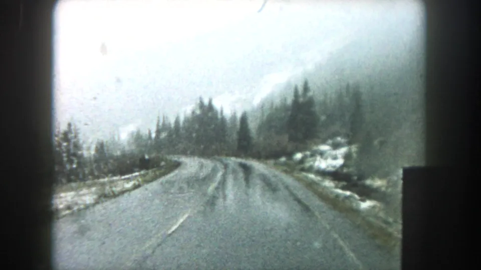 Forward car view on a wet winding road in Summit County, Colorado, bordered by evergreens, snow patches, and misty slopes.