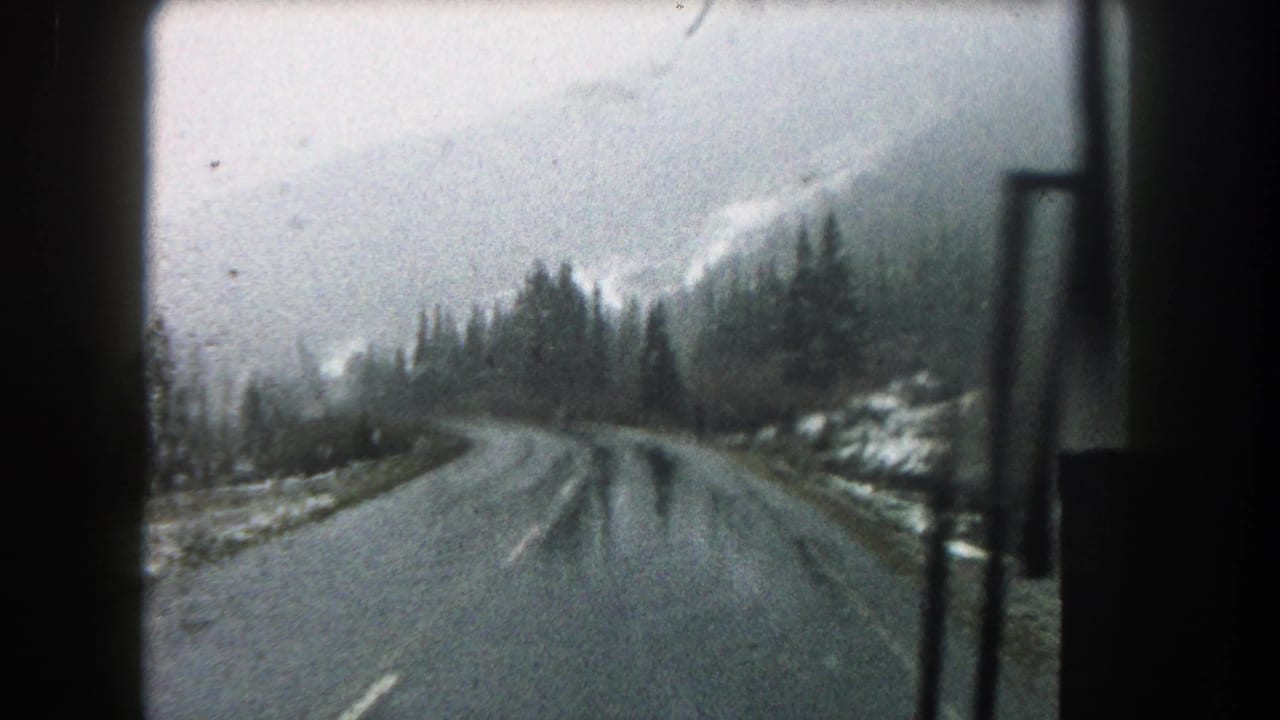1962 8mm view from inside a vehicle on a rainy Summit County mountain road with trees, mist, and windshield framing.