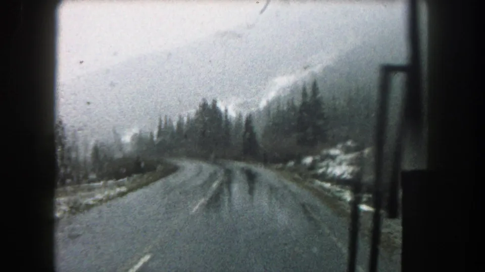 1962 8mm view from inside a vehicle on a rainy Summit County mountain road with trees, mist, and windshield framing.