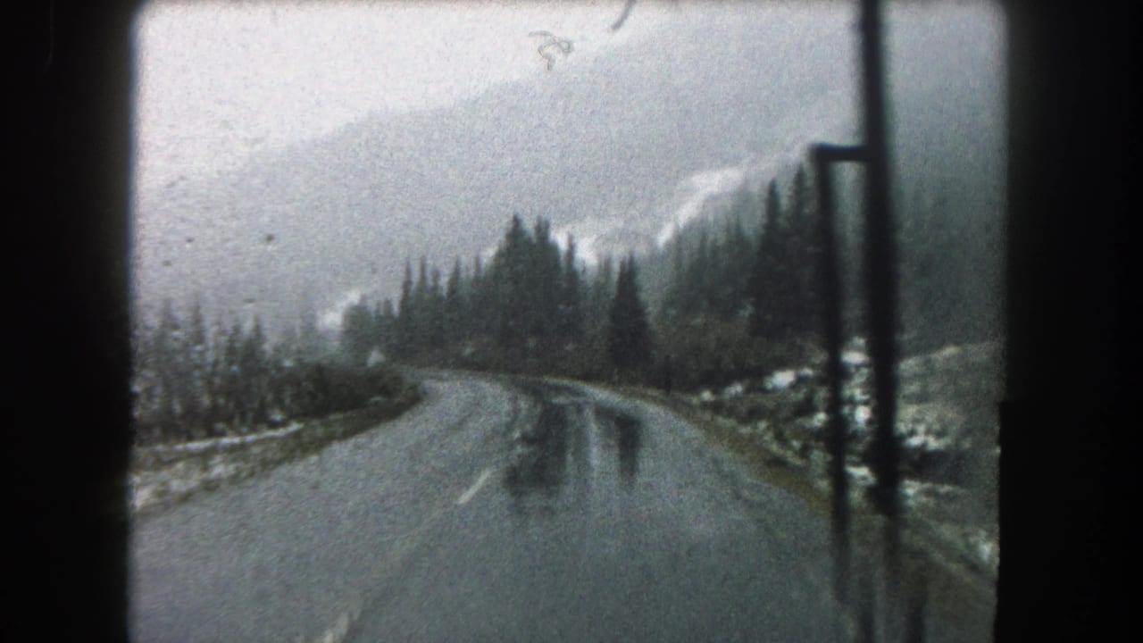 Vehicle-window view of a wet Summit County road in 1962, with evergreen trees, misted peaks, and dark interior framing.