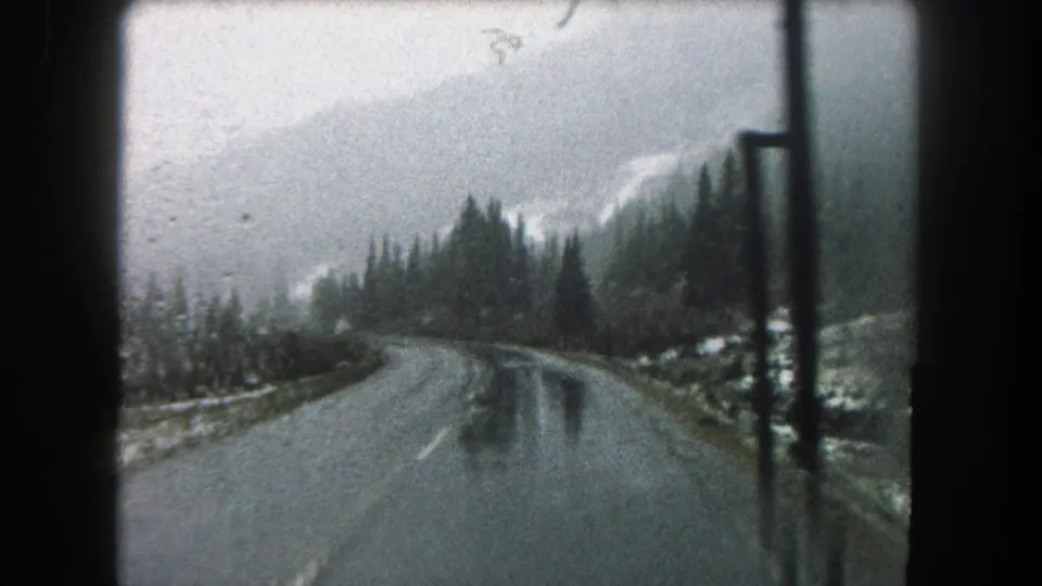 Vehicle-window view of a wet Summit County road in 1962, with evergreen trees, misted peaks, and dark interior framing.