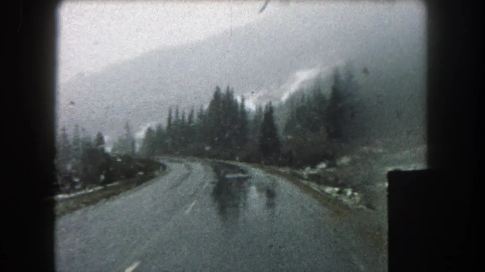 Soft 8mm frame of a rainy Summit County road, with the vehicle view looking toward evergreens and clouded mountain slopes.