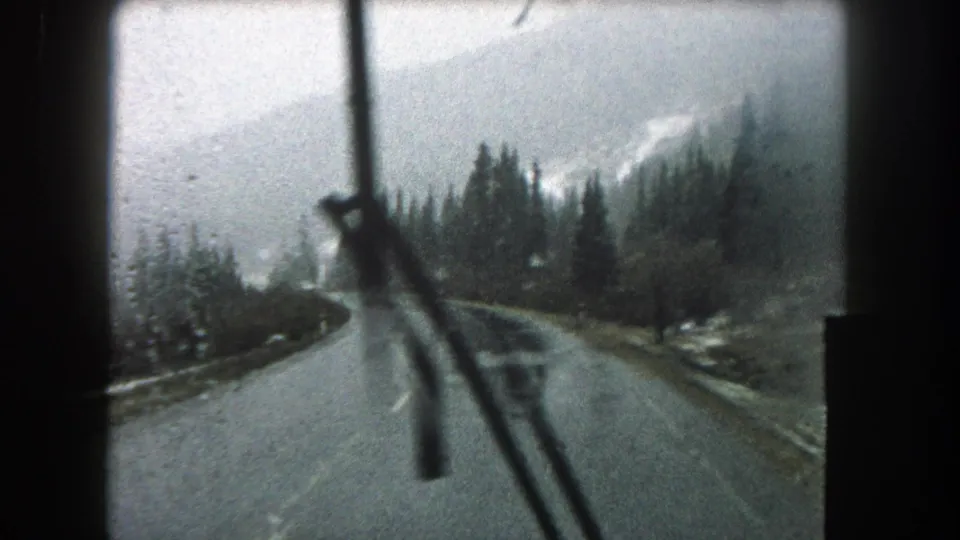 A windshield wiper sweeps across the 1962 Summit County road view, with wet pavement, trees, and misty mountains ahead.