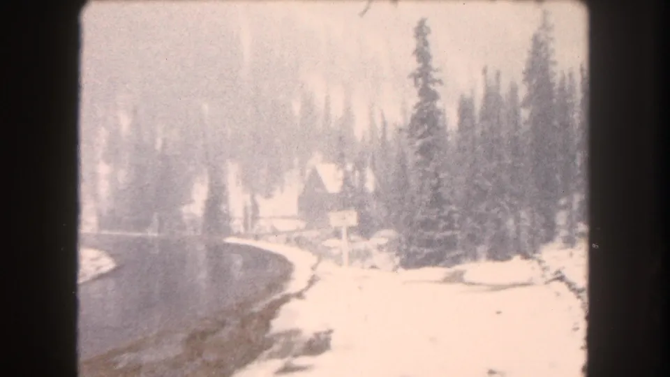 Passenger-view 8mm frame of a snowy Colorado mountain road curving past evergreen trees and a small lodge-like building in the distance.