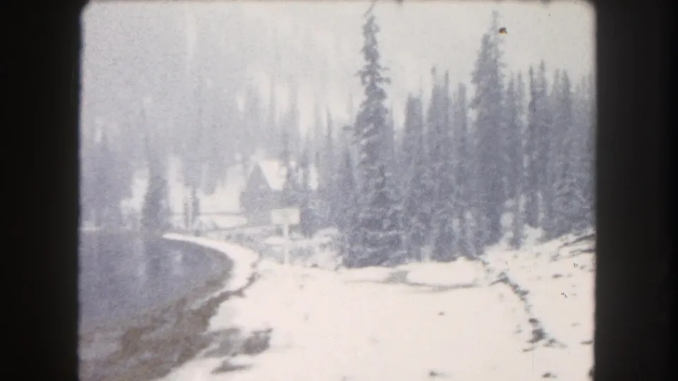 1962 Summit County, Colorado home-movie frame with a snowy lake edge, evergreens, and a small distant structure.