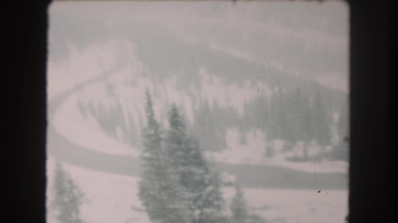 1962 8mm home-movie frame from Summit County, Colorado, showing snow-covered evergreens and a winding road below wooded slopes.