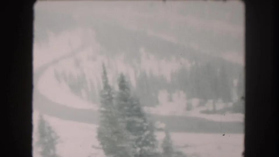1962 8mm home-movie frame from Summit County, Colorado, showing snow-covered evergreens and a winding road below wooded slopes.