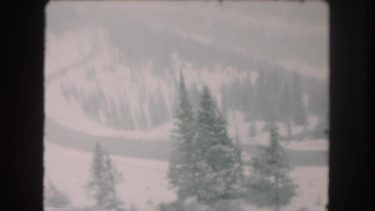 Soft 1962 8mm view of Summit County winter terrain, with evergreens, a winding road, and snowy wooded hills in the background.