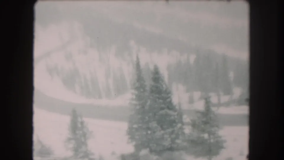 Soft 1962 8mm view of Summit County winter terrain, with evergreens, a winding road, and snowy wooded hills in the background.