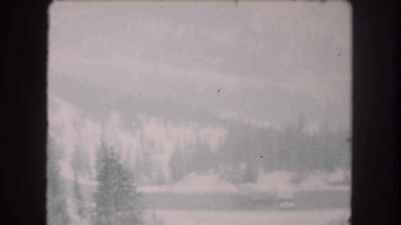 1962 Summit County, Colorado 8mm frame showing snow-covered terrain, dark evergreen trees, and distant wooded slopes under haze.