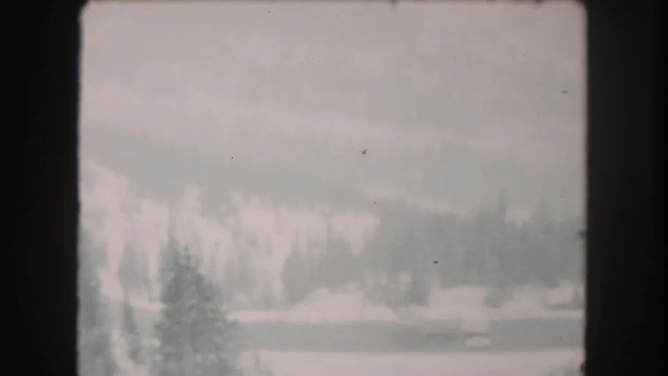 1962 Summit County, Colorado 8mm frame showing snow-covered terrain, dark evergreen trees, and distant wooded slopes under haze.