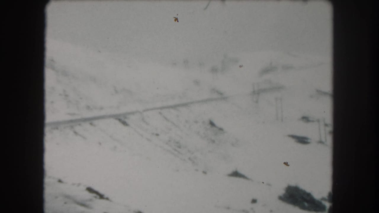 Snowy mountainside road with faint utility poles and dark 8mm film borders.