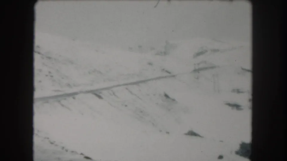 1962 Summit County, Colorado frame of a winter mountain road cutting across a pale snowy slope with distant poles.