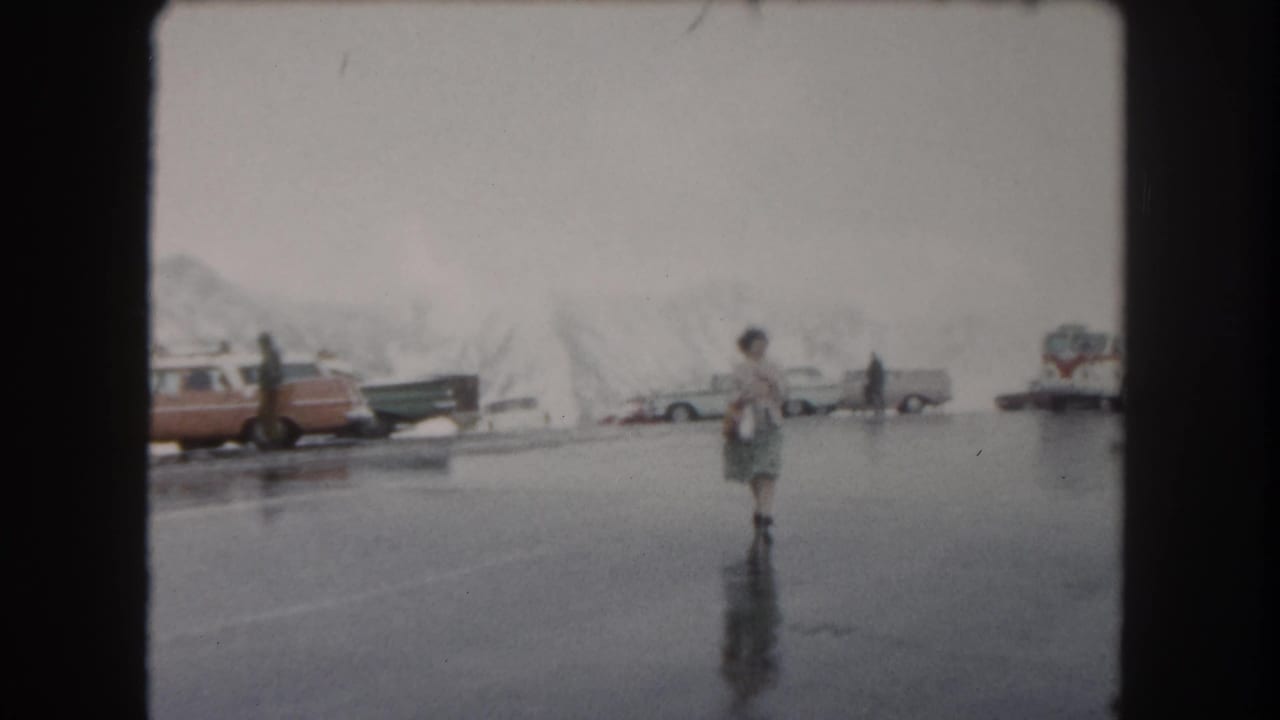 In 1962 Summit County, Colorado footage, a woman crosses reflective wet asphalt with snowy piles, parked cars, and a bus behind her.