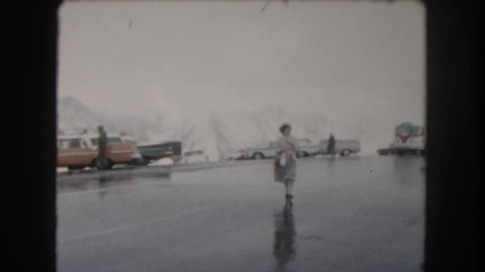 In 1962 Summit County, Colorado footage, a woman crosses reflective wet asphalt with snowy piles, parked cars, and a bus behind her.