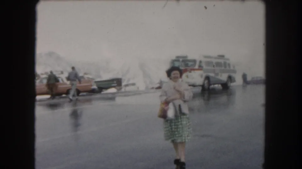 Near the end of the 1962 Summit County clip, the woman is close to camera with a bus, parked cars, pedestrians, and snowy piles behind her.