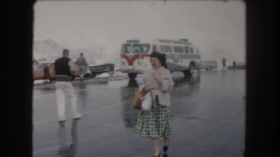 In 1962 Summit County, Colorado footage, a woman walks across wet pavement near a tour bus and parked cars.