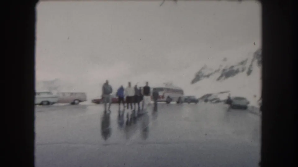 A distant group stands in a wet Summit County, Colorado parking area in 1962, with vintage cars and mountain slopes beyond.