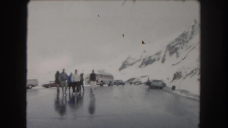 1962 Summit County, Colorado home-movie frame showing people walking across slick pavement below snow-covered mountain slopes.