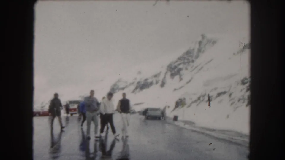 Mid-century home-movie frame of travelers crossing slick pavement in snowy Summit County, Colorado, with vehicles behind them.