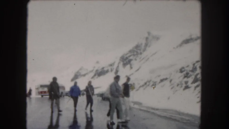 1962 Colorado mountain roadside scene with people standing and walking on reflective pavement near snowbanks and parked cars.