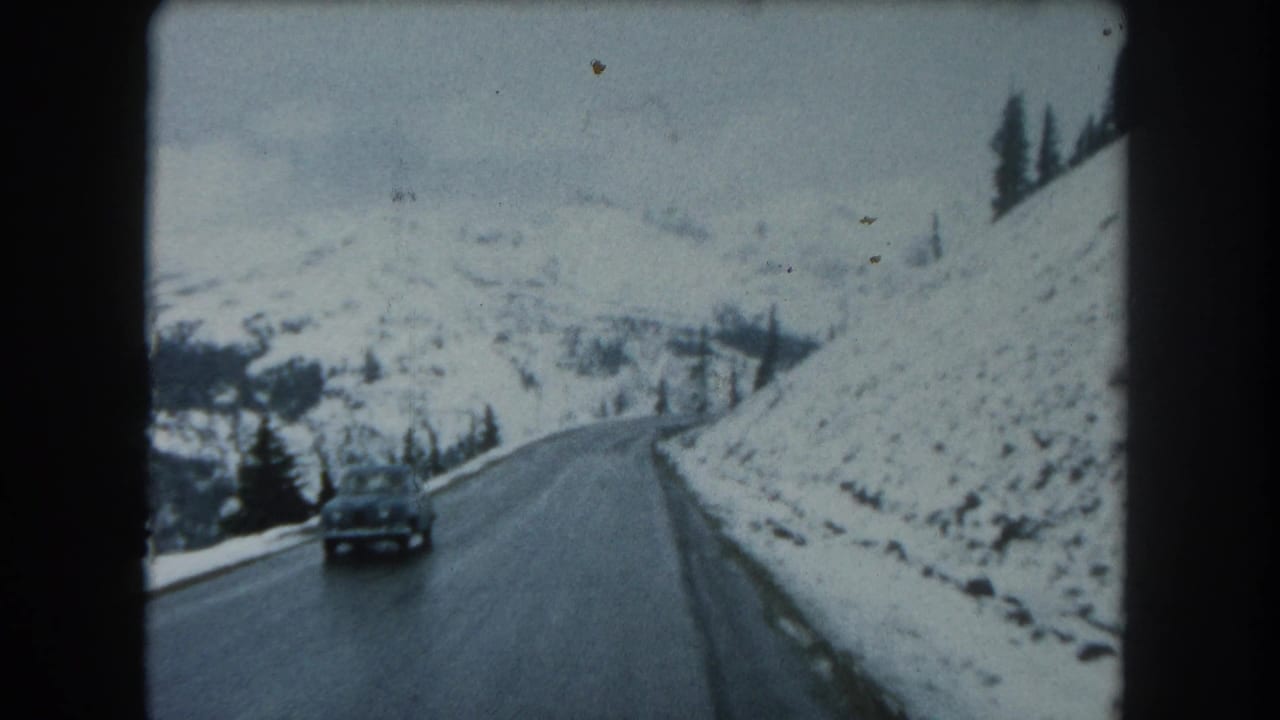 A dark car drives along a winding road through snow-covered mountains.