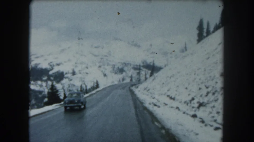 A vintage car approaches on a curving snowy mountain road in Summit County, Colorado, with evergreens and snowbanks along the route.
