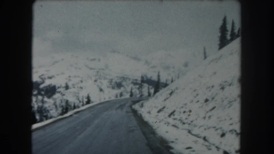 A vehicle-mounted or passenger viewpoint follows a winding wet road through snowy Summit County mountain terrain and sparse evergreens.