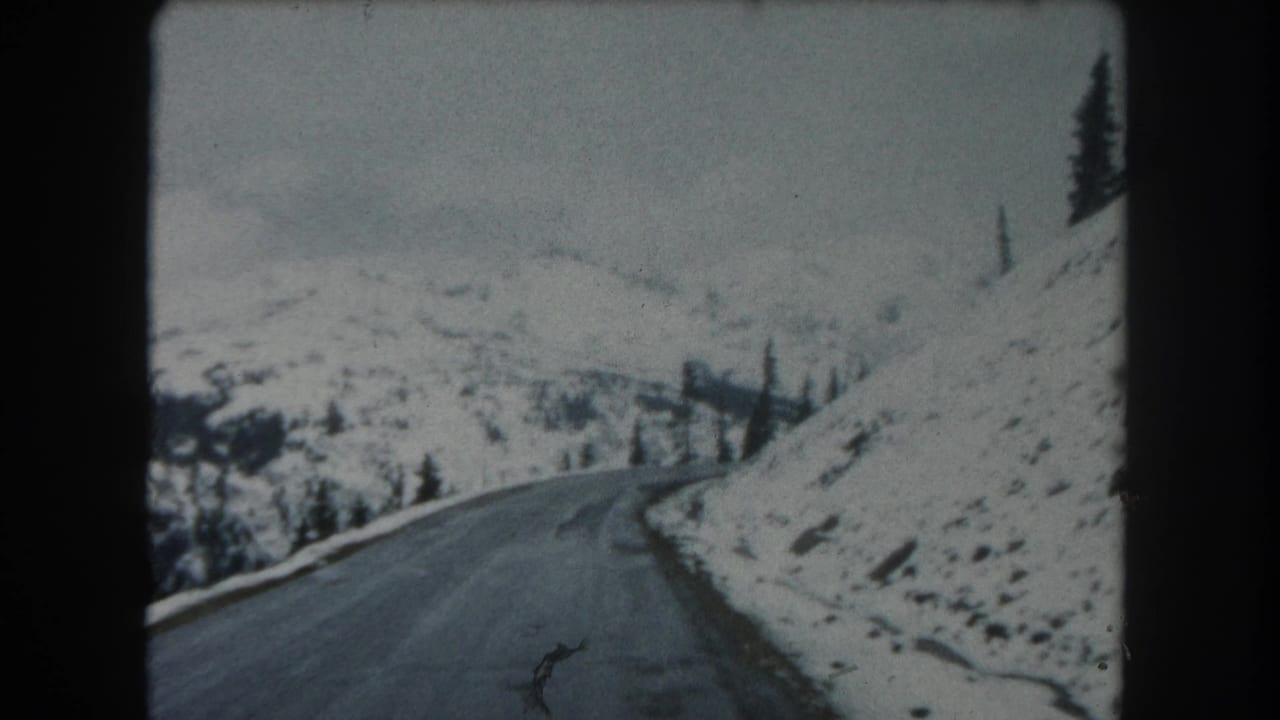 1962 Summit County, Colorado 8mm frame showing a snowy mountain road curving along a slope with evergreens and distant terrain.