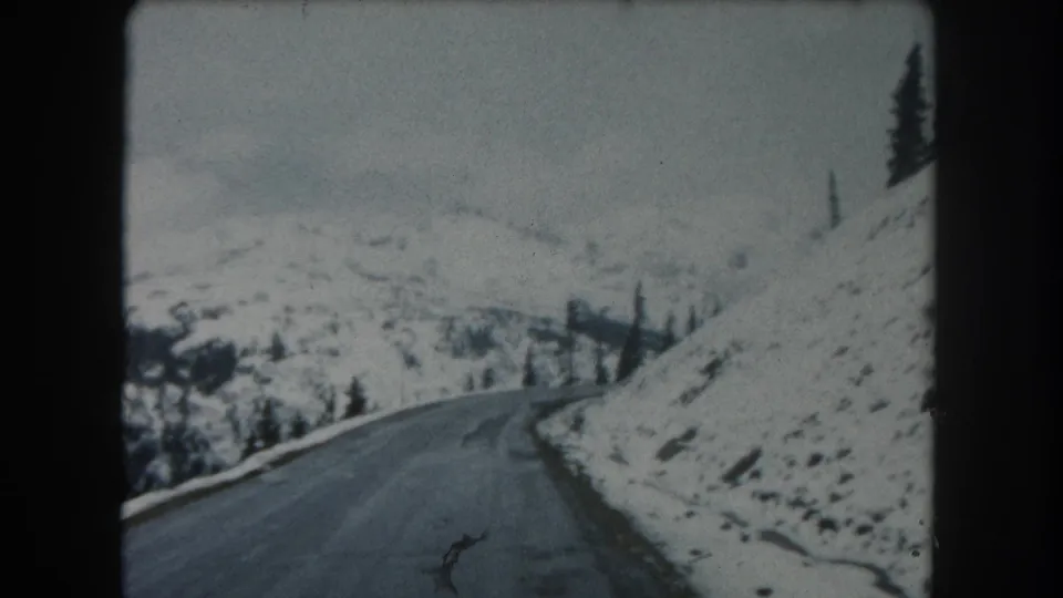 1962 Summit County, Colorado 8mm frame showing a snowy mountain road curving along a slope with evergreens and distant terrain.