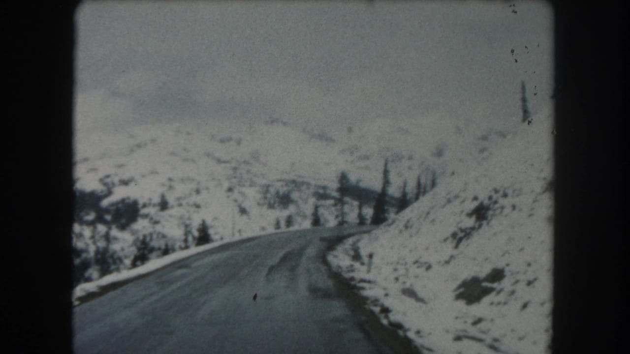 1962 Summit County, Colorado home-movie frame looking forward on a snowy mountain road with a right-hand bend and scattered evergreens.