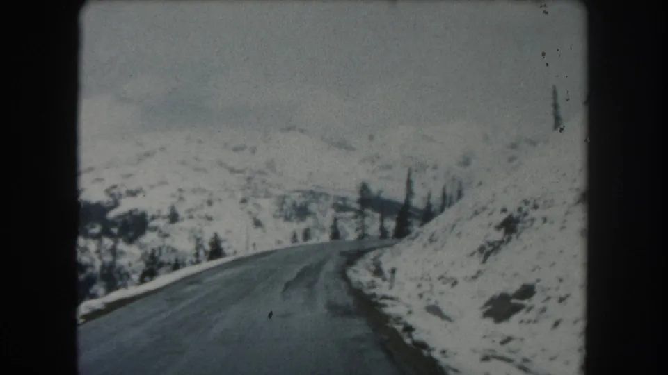 1962 Summit County, Colorado home-movie frame looking forward on a snowy mountain road with a right-hand bend and scattered evergreens.