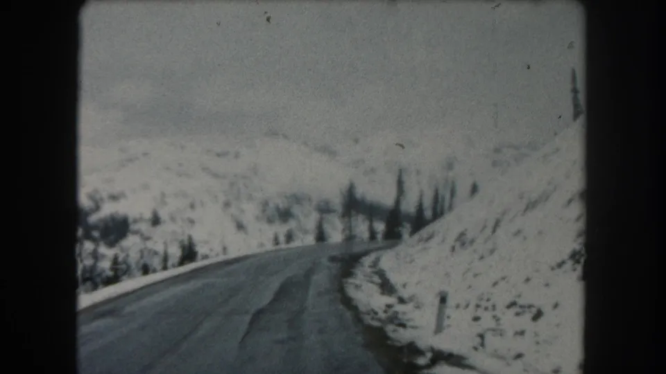 1962 Summit County, Colorado 8mm frame from a moving vehicle on a mountain road bordered by snow and sparse trees.