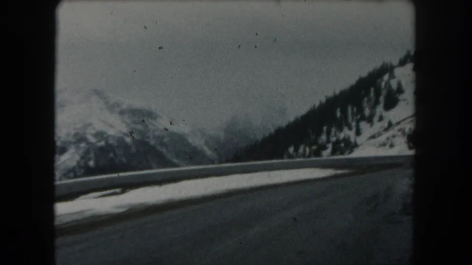 A moving-vehicle 8mm view shows a dark winter road edge, snow-covered mountains, and a tree-lined slope in Summit County, Colorado.