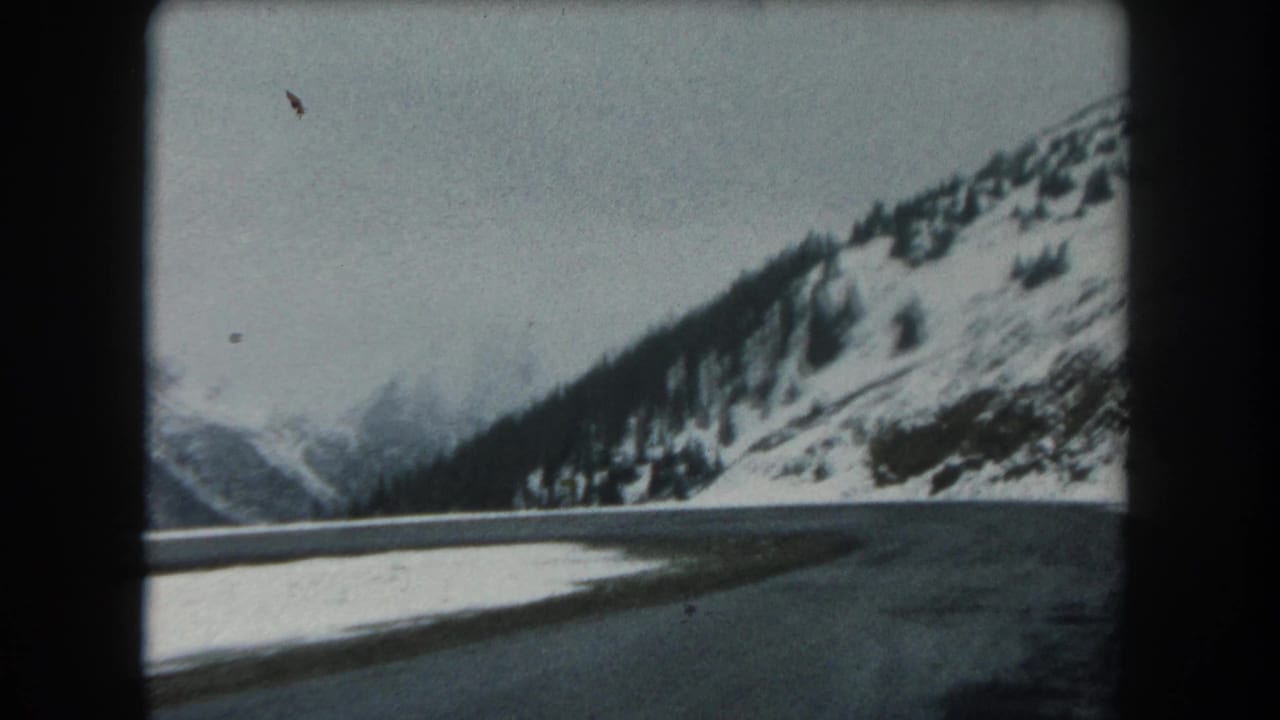 1962 8mm Summit County, Colorado view from a moving vehicle on a snowy mountain road with evergreen-covered slopes.