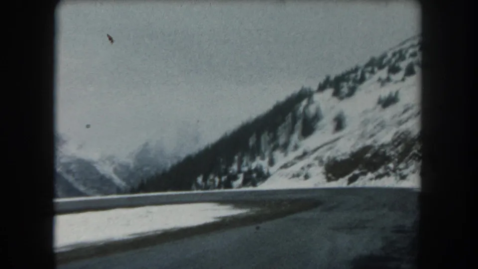 1962 8mm Summit County, Colorado view from a moving vehicle on a snowy mountain road with evergreen-covered slopes.