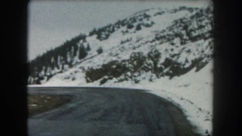 Vehicle-perspective 1962 8mm frame showing a Summit County road curve beside snowy Colorado mountain terrain.