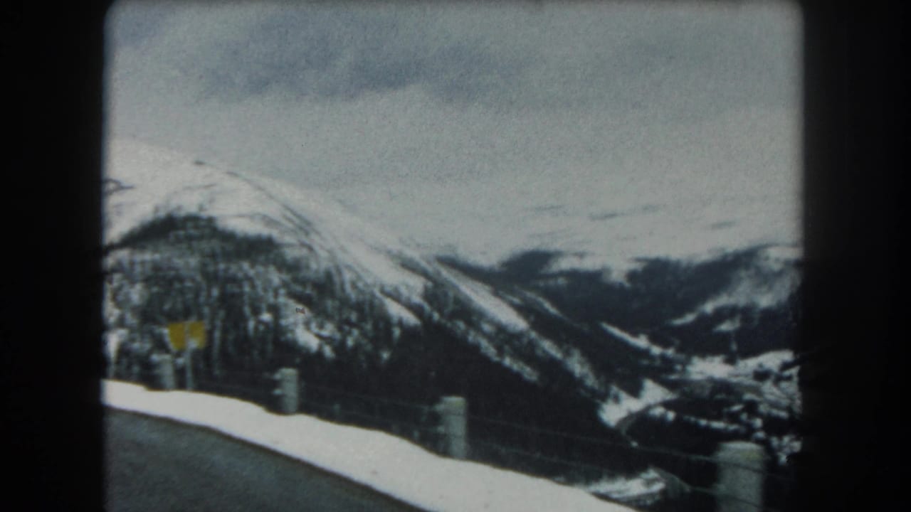 1962 Summit County, Colorado road-view frame with guardrail posts, a small yellow sign, snowy peaks, and wooded slopes.
