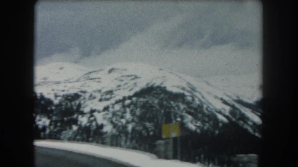 1962 Summit County, Colorado 8mm frame with a snowy ridge, muted sky, roadside sign, and foreground road edge.