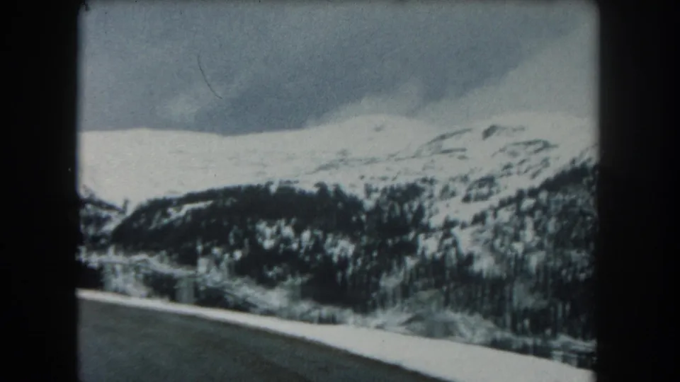 1962 Summit County, Colorado frame showing a curving snowy roadside view toward forested slopes and pale mountain ridgelines.