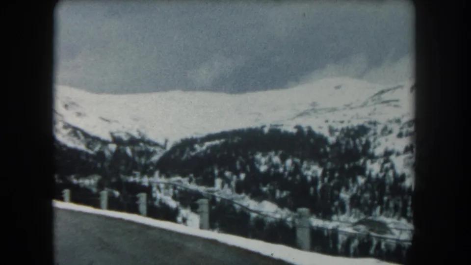 1962 Summit County, Colorado 8mm frame showing a snowy roadside, dark forested slopes, and distant white mountain ridges under cloud.