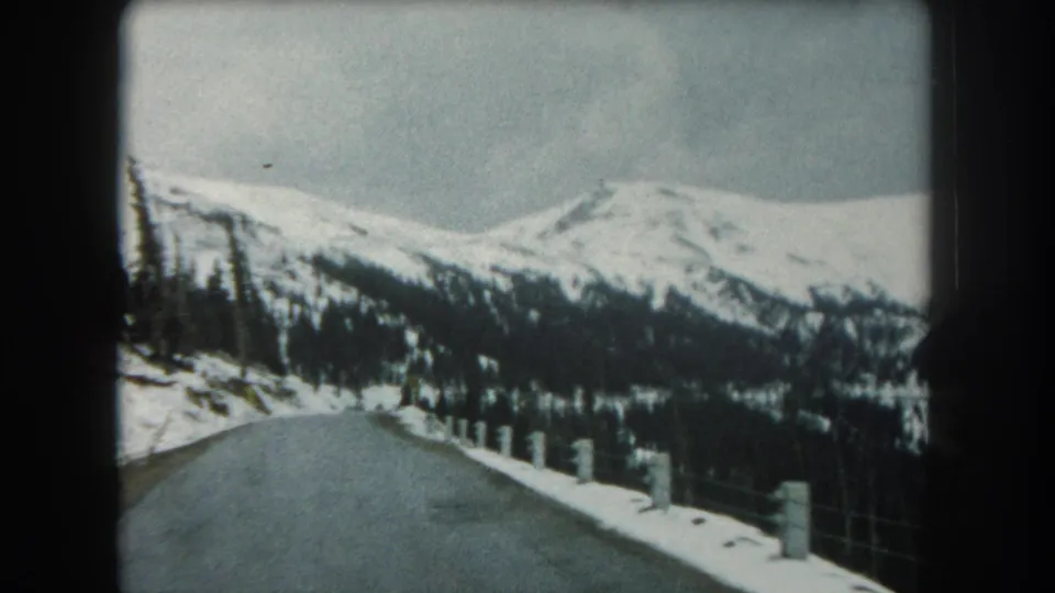 1962 Summit County, Colorado 8mm view down a winter mountain road bordered by snowbanks, roadside posts, and evergreen forest.
