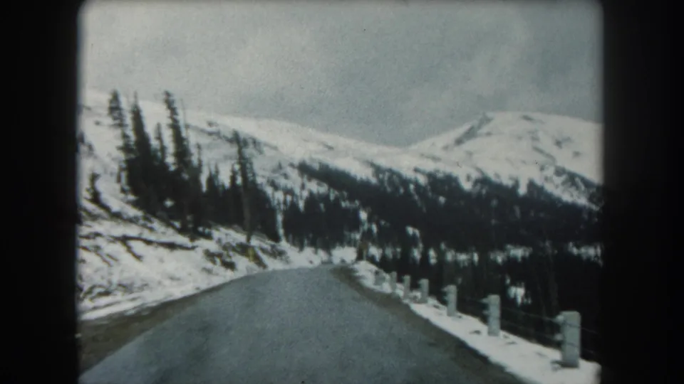 1962 Summit County, Colorado frame showing a snow-lined road, sparse roadside posts, evergreen trees, and pale mountain slopes ahead.