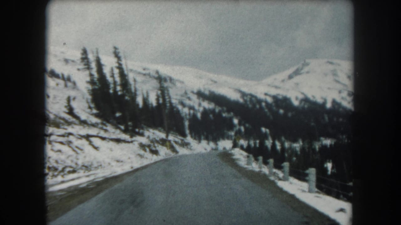 Snowy mountain road curving through evergreen forest toward distant ridges.